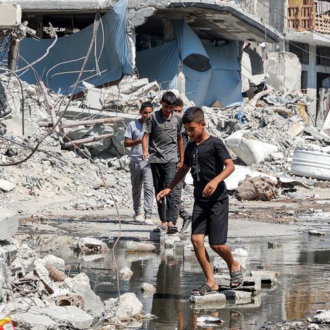 Boys walk on dry bricks to traverse a puddle of sewage water past mounds of trash and rubble along a street in the Jabalia camp for Palestinian refugees, northern Gaza Strip, August 14, 2024.